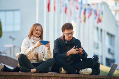 Akıllı telefonu tutuyorsun. İki genç öğrenci dışarıda üniversiteye karşı oturuyorlar..