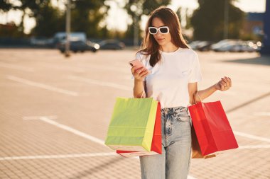 Akıllı telefon kullanıyor. Günlük giysiler içinde güzel bir kadın alışveriş torbaları tutuyor, açık havada..