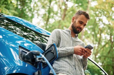 Aşağıdan bak, akıllı telefonla. Yakışıklı adam dışarıda elektrikli otomobilin yanında duruyor ve şarj edilmeyi bekliyor..