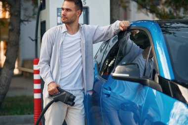 Elinde kabloyla. Şarj istasyonunda mavi elektrikli arabası olan adam..