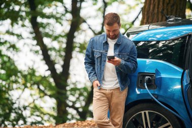 Ayakta ve akıllı telefonu tutarak. Yakışıklı adam dışarıda elektrikli arabasının şarj olmasını bekliyor..