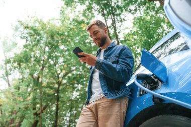 Aşağıdan bak, akıllı telefonla. Yakışıklı adam dışarıda elektrikli arabasının şarj olmasını bekliyor..
