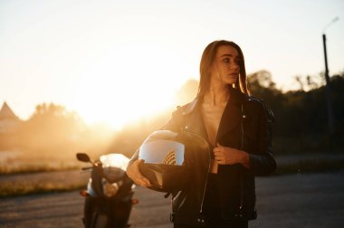 Koruyucu miğferi elinde tutuyor. Dışarıda motosikletli bir kadın var..