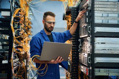 Dizüstü bilgisayarı kullanarak. Genç adam sunucu odasında internet ekipmanı ve kablolarıyla çalışıyor..