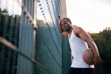 Metal yapıya yaslanarak. Genç siyah adam basketbol topuyla dışarıda..