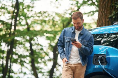 Ayakta ve akıllı telefonu tutarak. Yakışıklı adam dışarıda elektrikli arabasının şarj olmasını bekliyor..