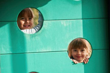 İki delikten bakıp gülümsüyor. Çocuklar oyun alanında eğleniyor..