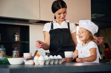 Kız şef şapkası takıyor. Anne ve kızı mutfakta yemek hazırlıyorlar..