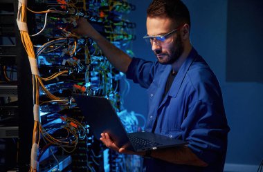 Gözlüklü. Genç adam sunucu odasında internet ekipmanı ve kablolarıyla çalışıyor..
