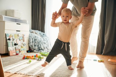 Oğlan babasının yardımıyla yürüyor. Adam evcil odasında oğluyla oynuyor..