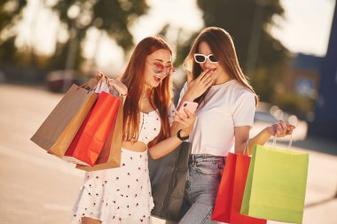 Akıllı telefondaki içerik beni heyecanlandırdı. Günlük giysiler içinde iki güzel kadın ellerinde alışveriş çantaları, açık havada..