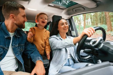 Gülümsüyor, eğleniyor. Baba, anne ve küçük oğlu modern arabada oturuyorlar..