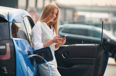 Akıllı telefon ellerde. Elektrikli arabalardaki kadın gündüz şarj istasyonunda.