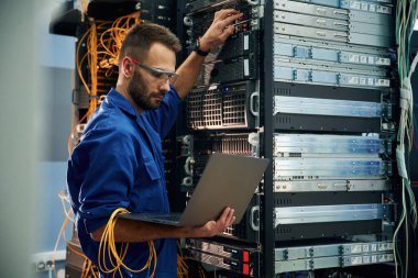 Dizüstü bilgisayarı kullanarak. Genç adam sunucu odasında internet ekipmanı ve kablolarıyla çalışıyor..