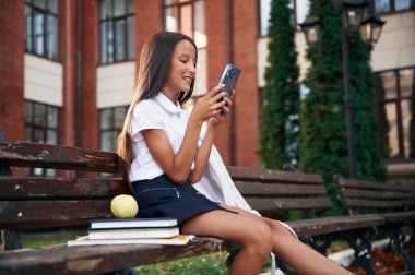 Akıllı telefonla bankta oturmak. Okul üniformalı kız binanın yanında..