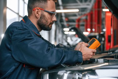 Elektriği ölçmek için sarı renkli cihaz ellerde. Araba tamircisi otomobille garajda..