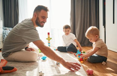 Güneş ışığı pencereden içeri giriyor. Babam yerde oyuncaklarla oynayan iki küçük çocukla oynuyor..
