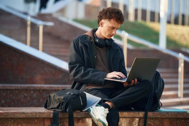 Adam oturuyor ve dizüstü bilgisayar kullanıyor. Genç öğrenci üniversitenin dışında, siyah elbiseli..