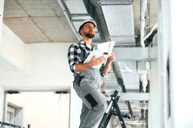 Evraklar ellerinde. Beyaz kasketli. Üniformalı tamirci tamamlanmamış binada..
