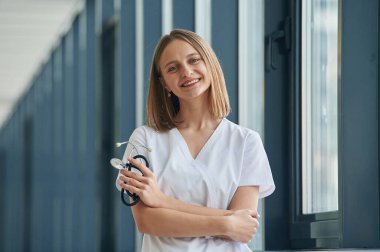 Stetoskop elinde, ayakta duruyor. Beyaz önlüklü kadın doktor koridorda..