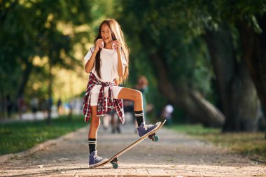 Arka planda, patikada yeşil ağaçlar var. Dışarıda kaykaylı mutlu küçük kız.