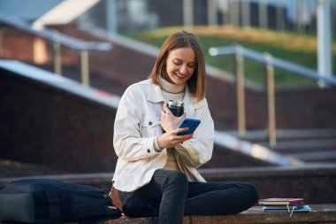 Akıllı telefon ellerde. Gündelik giysiler içinde genç bir kız öğrenci dışarıda..