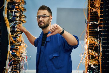 Baş parmağını aşağı gösteriyor. Genç adam sunucu odasında internet ekipmanı ve kablolarıyla çalışıyor..