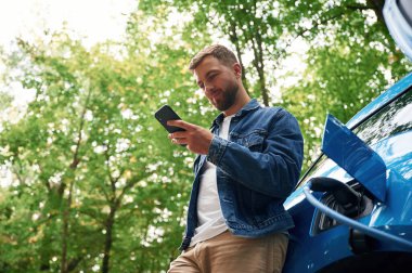Aşağıdan bak, akıllı telefonla. Yakışıklı adam dışarıda elektrikli arabasının şarj olmasını bekliyor..