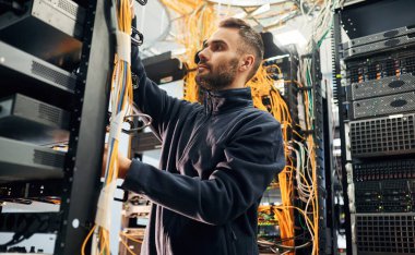 Siyah kıyafetler. Genç adam sunucu odasında internet ekipmanı ve kablolarıyla çalışıyor..