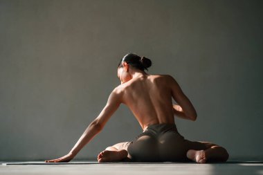 Sütyensiz, yerde oturuyor. Zayıf vücutlu genç bir kadın stüdyoda spor kıyafetleri giyiyor..