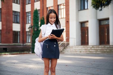 Kablosuz cihaz elinde, ayakta duruyor. Okul üniformalı kız binanın yanında..