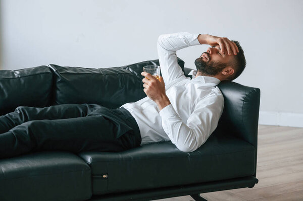 Голова болит, лежит на диване. Расстроенный молодой человек пьет алкоголь в помещении.