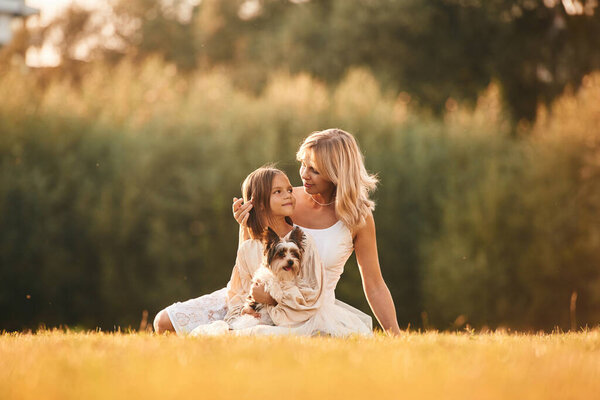 Sitting on the ground. Mother with her daughter and cute dog are on the field outdoors.