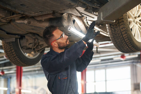 In blue colored uniform. Car repairman is in the garage with automobile.