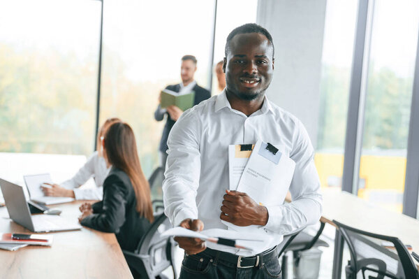 Black man is standing and holding documents. Group of office workers are together indoors.