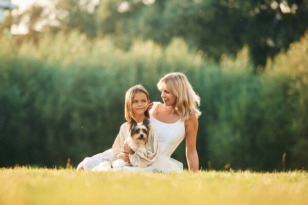 Sitting on the ground. Mother with her daughter and cute dog are on the field outdoors.