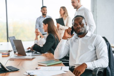 Siyah adam telefonun yanında konuşuyor. Bir grup ofis çalışanı içeride birlikte..