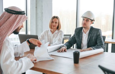 Masanın yanında bir mimar oturuyor. Geleneksel kıyafetli Müslüman işadamı ve görevde meslektaşları.