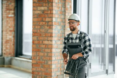 Ellerinde siyah kablolar var. Bitmemiş odada üniformalı inşaat işçisi..