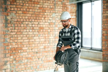 Ellerinde siyah kablolar var. Bitmemiş odada üniformalı inşaat işçisi..