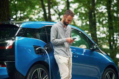 Kablosuz cihaz, akıllı telefon kullanıyor. Yakışıklı adam dışarıda elektrikli otomobilin yanında duruyor ve şarj edilmeyi bekliyor..