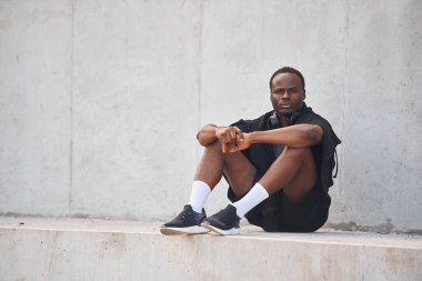 Beyaz duvarın yanında oturuyor. Yakışıklı siyah adam gündüz vakti dışarıda poz veriyor..