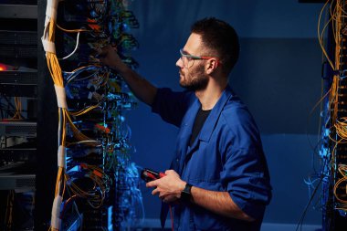 Mavi renkli ışıklandırma. Genç adam sunucu odasında internet ekipmanı ve kablolarıyla çalışıyor..