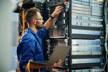 Dizüstü bilgisayarı kullanarak. Genç adam sunucu odasında internet ekipmanı ve kablolarıyla çalışıyor..