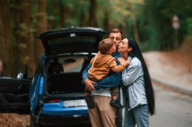 Baba, anne ve oğul dışarıda mavi arabanın yanında eğleniyorlar..