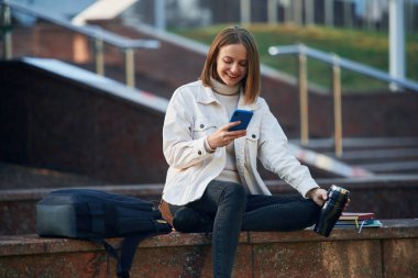 Akıllı telefon ellerde. Gündelik giysiler içinde genç bir kız öğrenci dışarıda..