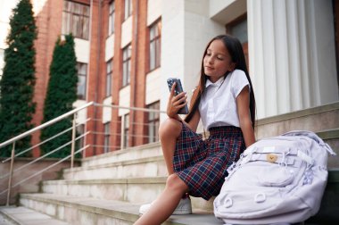 Merdivenlerde oturup, akıllı telefon kullanmak. Okul üniformalı kız binanın yanında..