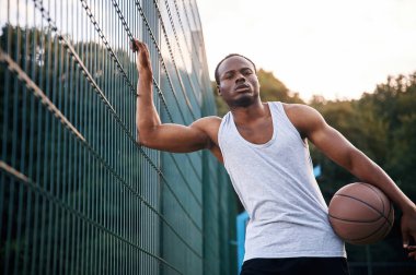 Metal yapıya yaslanarak. Genç siyah adam basketbol topuyla dışarıda..