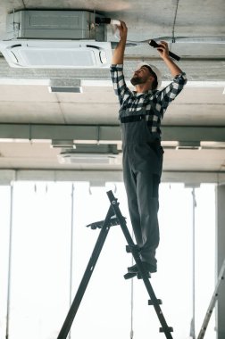 Merdivenin üstünde duruyor. Üniformalı tamirci tamamlanmamış binada..