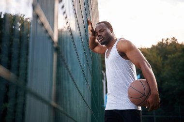 Metal yapıya yaslanarak. Genç siyah adam basketbol topuyla dışarıda..